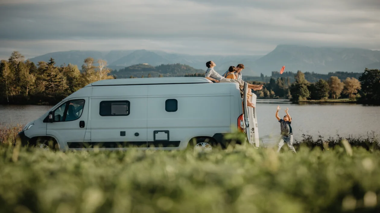 Caravan with three people on top and green surroundings with a blue sky.