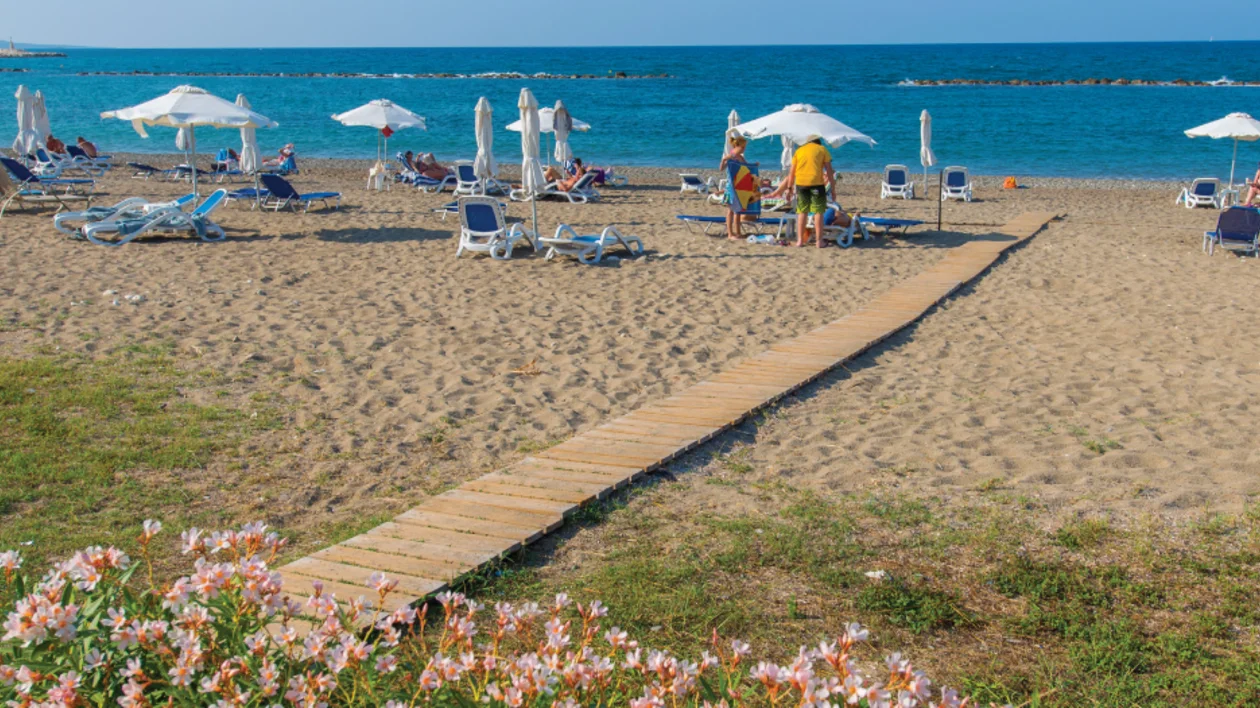 Beach with lounge chairs and umbrellas along a wooden walkway, overlooking a calm blue sea and distant hills.