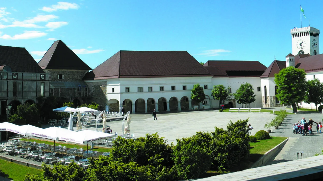 A castle and its clock tower, with a square with a lively café, trees and benches.