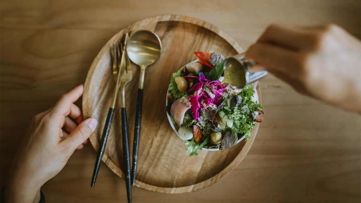 A vibrant salad with various vegetables and colourful garnishes, served on a wooden plate, with a hand pouring dressing over it.