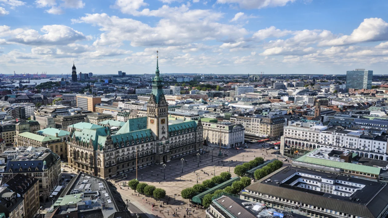 A stunning panoramic view of Hamburg’s city centre, with the magnificent Hamburg Rathaus (city hall) as the focal point. The surrounding buildings, green roofs, and the distant harbour create a blend of historic and modern architecture, all under a bright, partly cloudy sky.