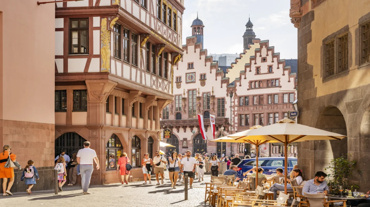 A bustling street scene in Frankfurt's old town, Römerberg. People stroll through the square near traditional timber-framed buildings, with outdoor seating from nearby cafes offering a perfect view of the historic architecture. The iconic Römer city hall is visible in the background.