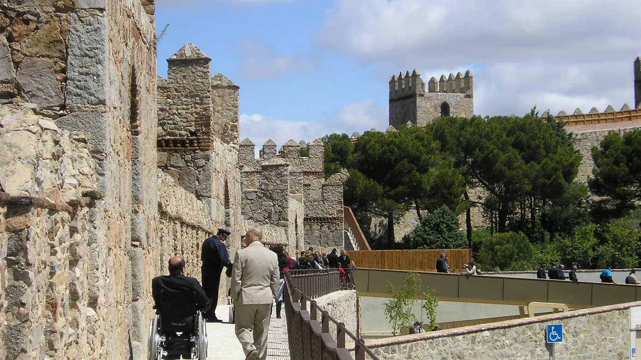 People walking near an ancient stone wall with towers under a clear sky. One of them is a wheelchair user.