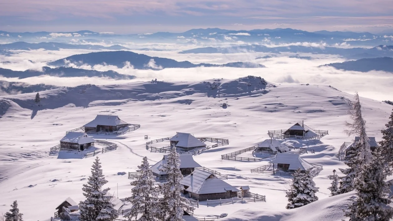 Snow-covered alpine village with wooden huts overlooking clouds and distant mountains.