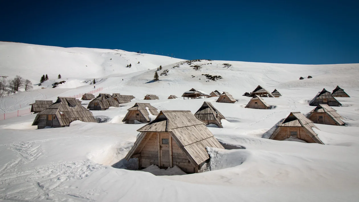 A village of traditional wooden huts partially buried in snow on a pristine white mountainside, under a clear blue sky.