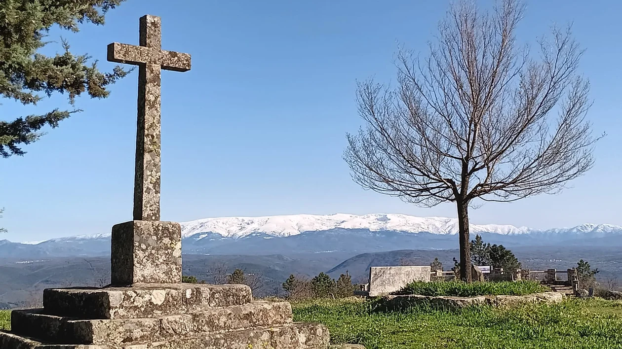 Stone cross and bare tree overlooking snowy mountains under a clear blue sky.