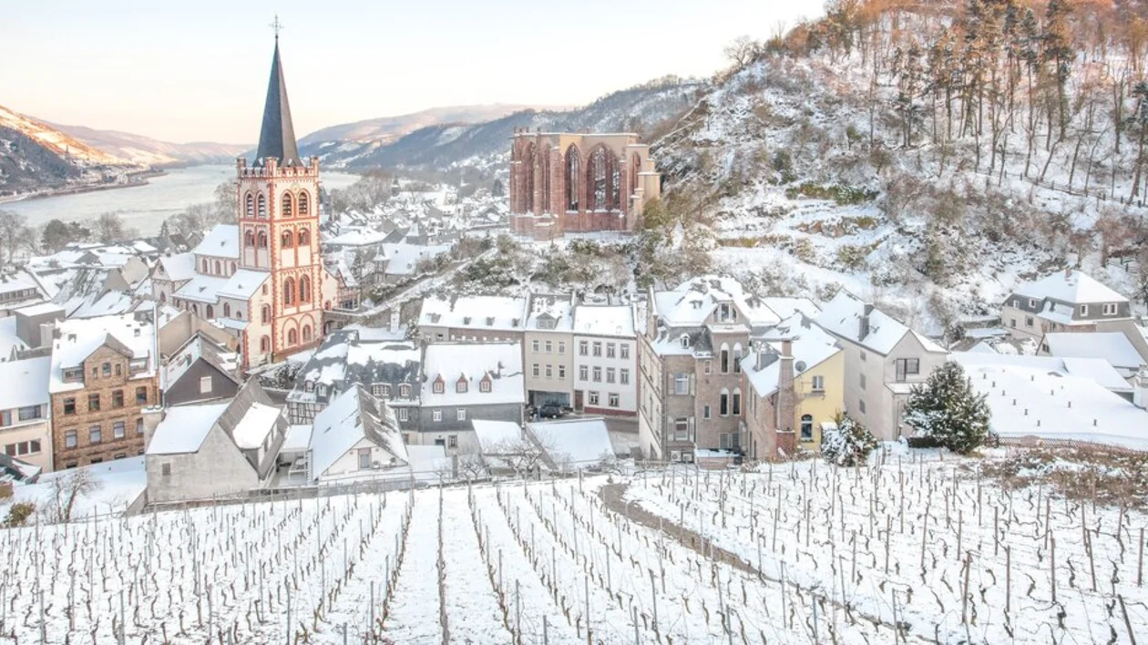 A snowy European village with a church, buildings, and vineyards by a river.