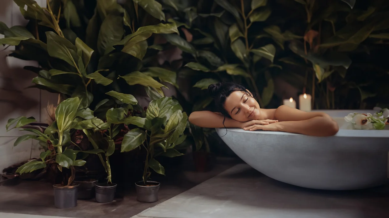 A woman relaxing in a bathtub surrounded by lush green plants and lit candles.