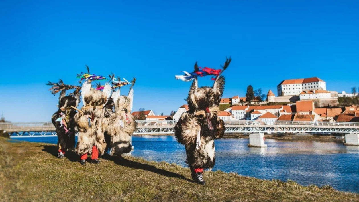 People in traditional costumes near a river with a bridge and town in background.
