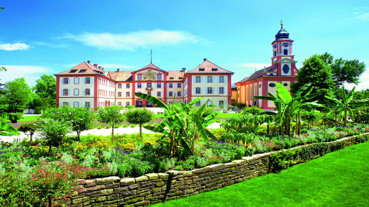 A daytime view of the Insel Mainau castle and gardens, featuring colorful flowers, trees, and elegant architecture.