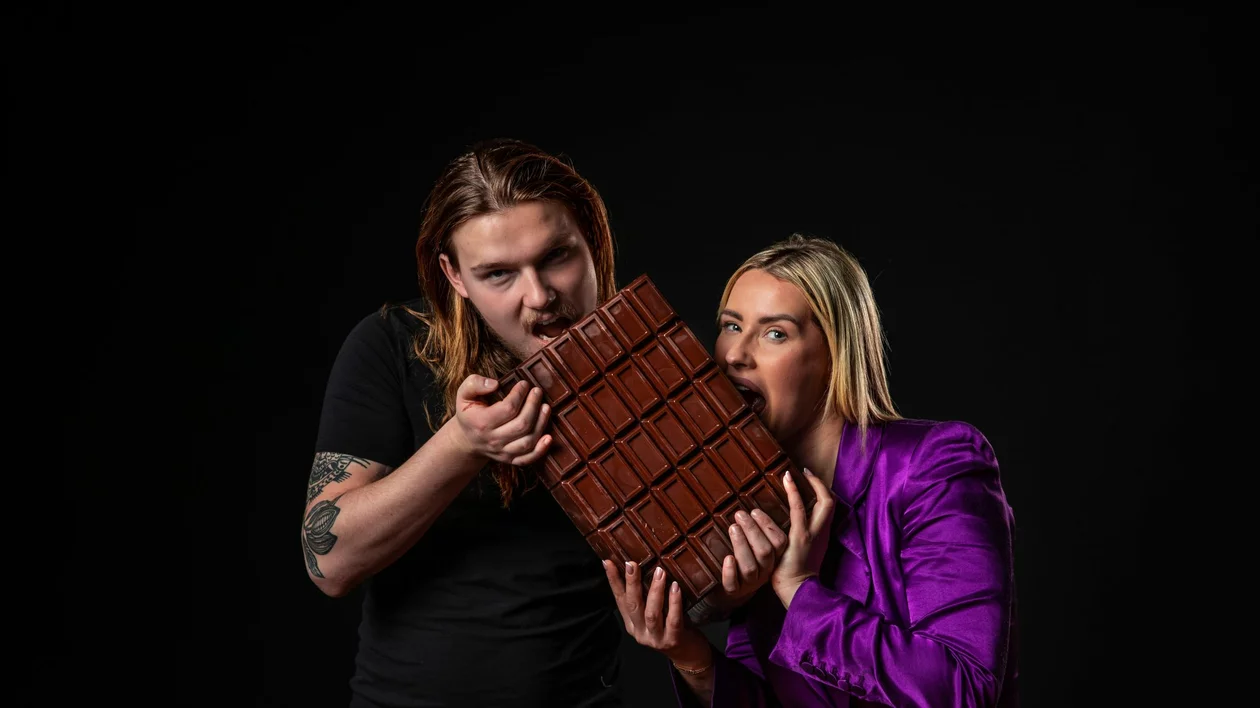 A man and woman hold a large chocolate bar, smiling and positioned against a dark background. The man has long hair and tattoos; the woman wears a purple blazer.