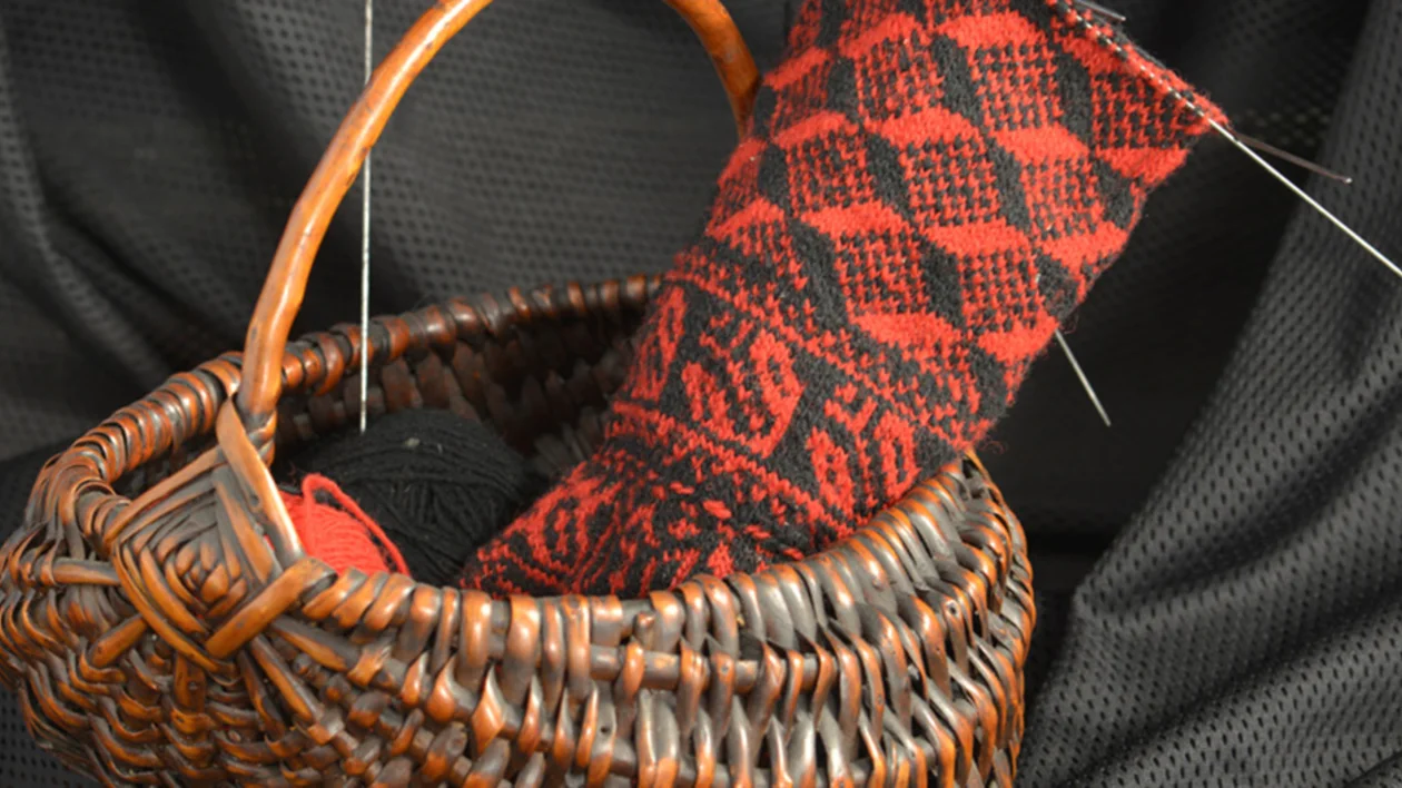 A woven basket holds a partially completed red and black patterned textile piece with sewing needles, against a dark fabric backdrop.