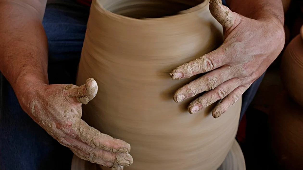 A potter shapes a spinning clay pot on a wheel, hands covered in wet clay, showcasing the artistry of ceramics.