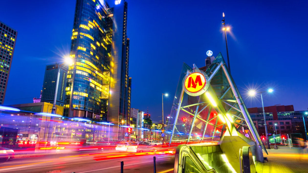 A modern metro station entrance in Warsaw illuminated at night, with skyscrapers and city traffic blurred in motion.