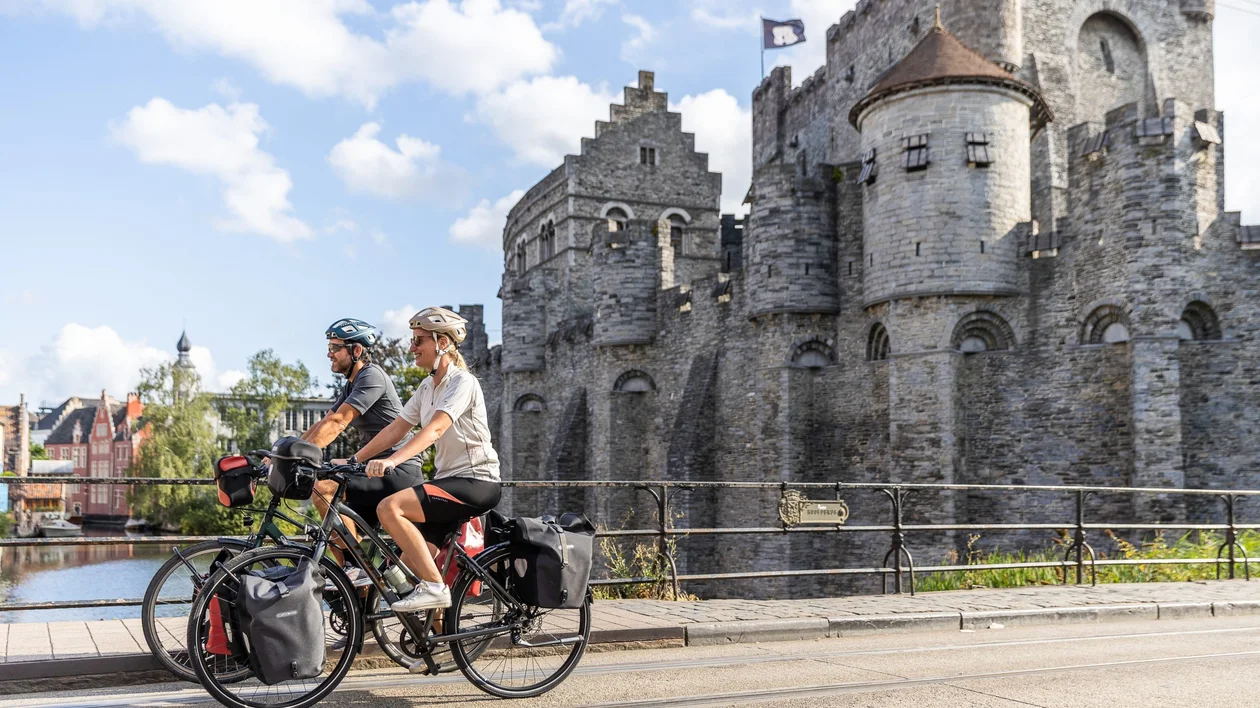 Two cyclists with touring bikes ride past Gravensteen Castle in Ghent, Belgium, with a blue sky and historic buildings in the background.