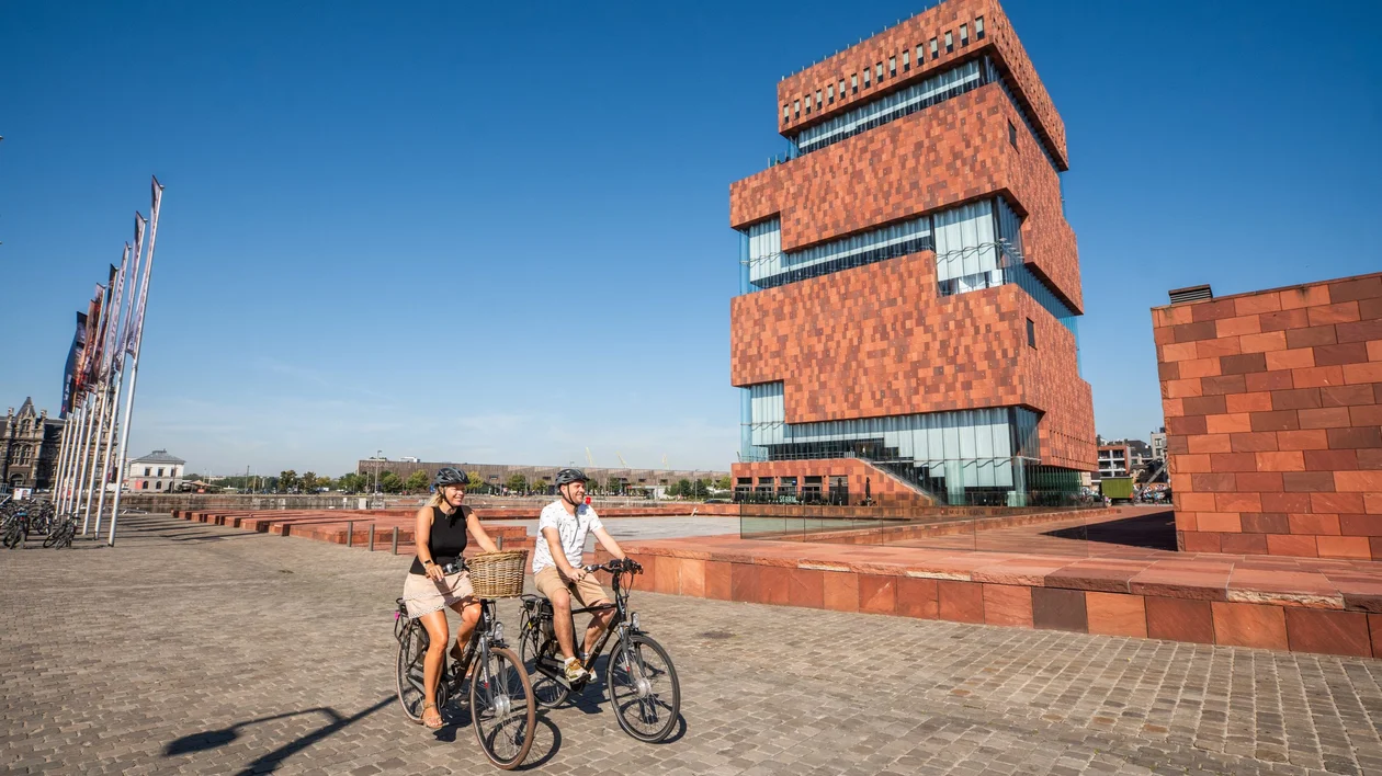 A couple rides bicycles on a cobblestone path in front of the MAS Museum in Antwerp, Belgium, on a sunny day.