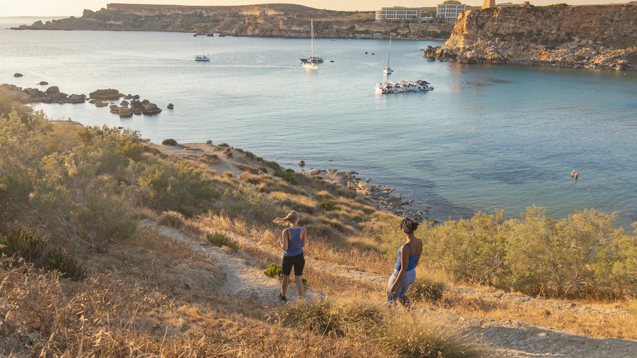 Two women hike down a grassy slope towards a serene bay, dotted with boats, under a warm sunset glow in Malta.