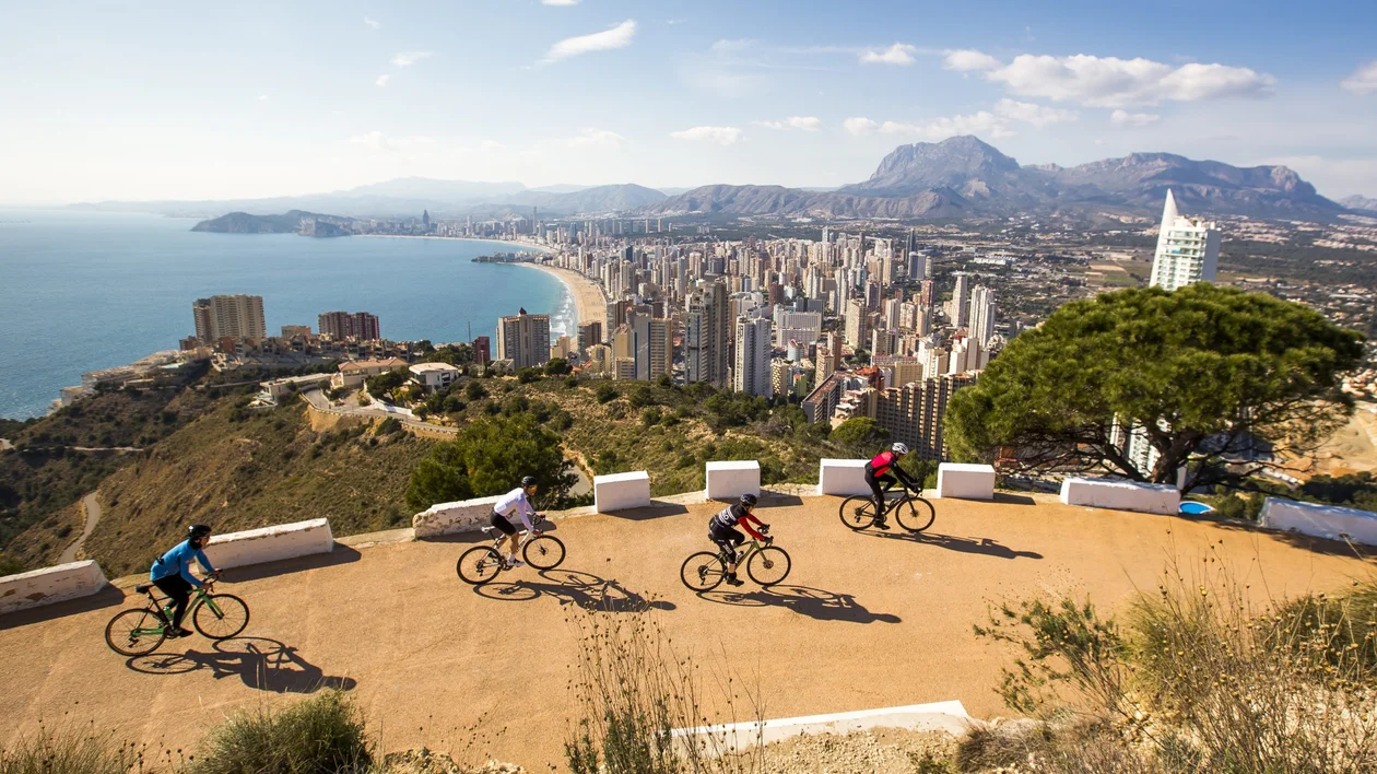 Group of cyclists riding along a scenic coastal road with a panoramic view of a city skyline, sandy beach, and mountainous landscape in the background. The bright, sunny day highlights the contrast between the blue sea and the rugged terrain.
