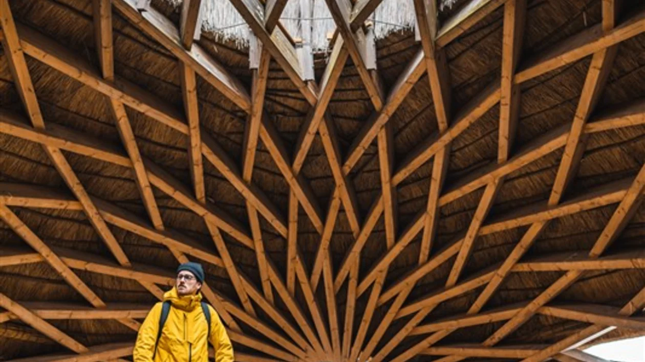 A person in a yellow jacket stands on a circular balcony beneath a striking wooden star-patterned ceiling with an opening at the center.
