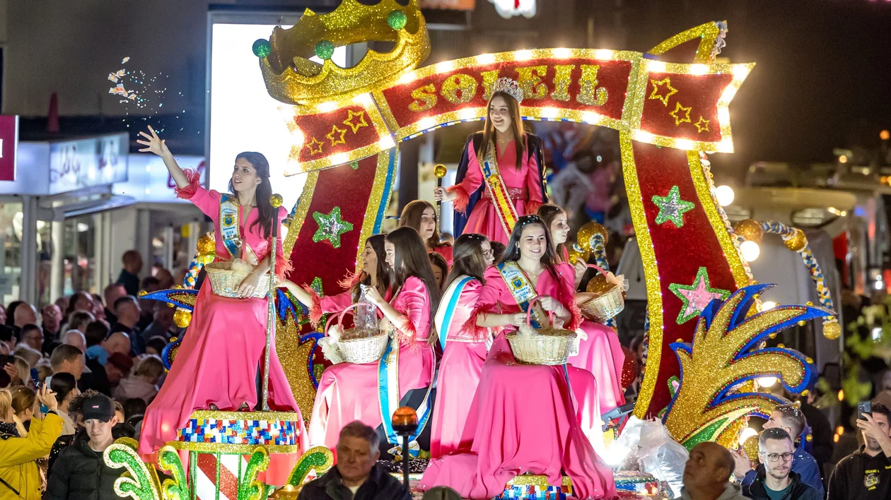 A festive parade float adorned with colorful decorations, featuring performers in pink dresses holding baskets, surrounded by an enthusiastic crowd.
