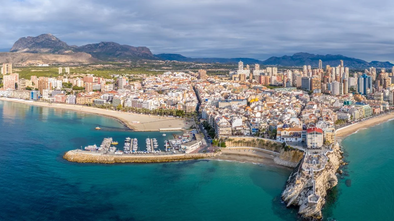 Aerial view of a coastal city with sandy beaches, marina, and modern buildings against a backdrop of mountains and cloudy skies.