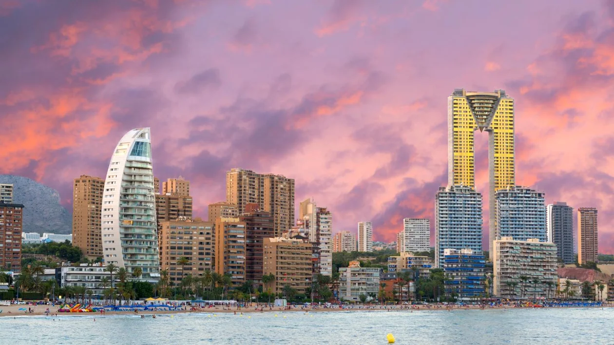 Vibrant sunset over a coastal city with modern buildings, palm trees, and a busy beach scene under colorful skies.