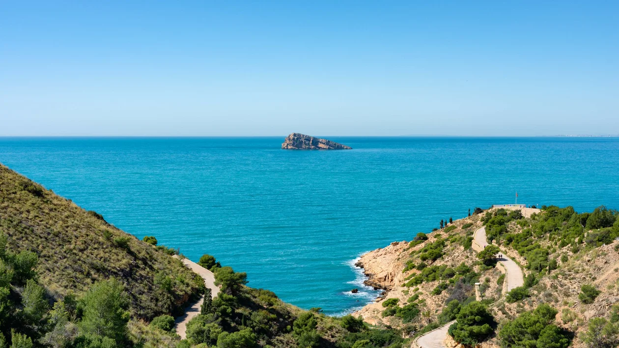 Serene coastal view featuring turquoise waters, rolling hills, and a distant rocky island under a clear blue sky.