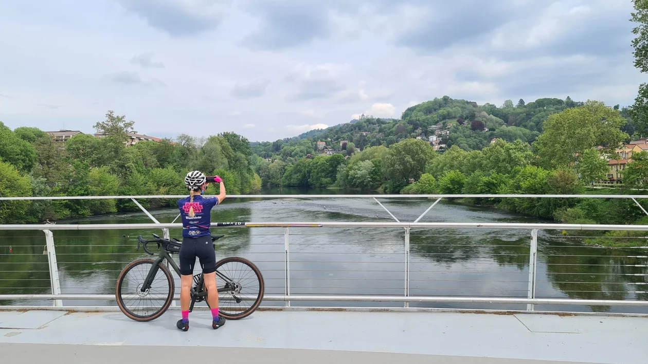 A cyclist in a blue jersey and pink accessories stands on a bridge, capturing the lush, green river landscape with a phone.