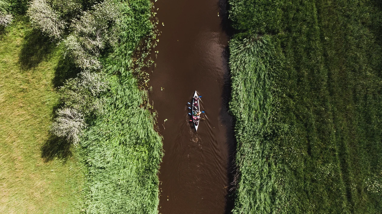 Aerial view of a canoe with several paddlers navigating a narrow, winding river bordered by lush greenery and wildflowers.