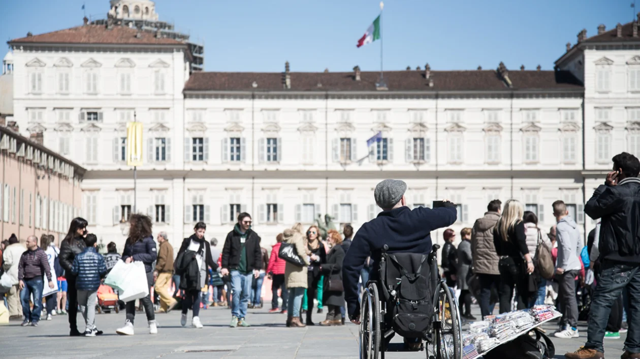 A man in a wheelchair takes a selfie amidst a busy plaza filled with people, with historic architecture in the background.