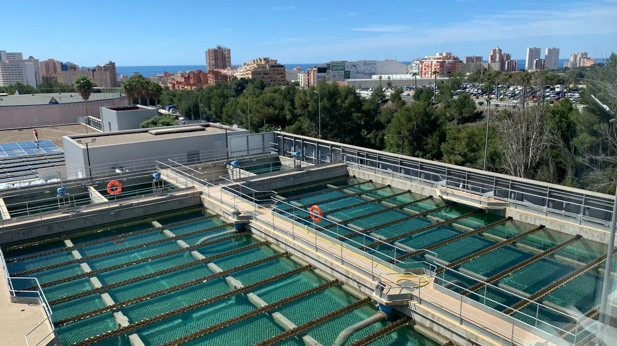 A view of a water treatment facility featuring large, green water tanks, adjacent to urban buildings and trees under a clear blue sky.