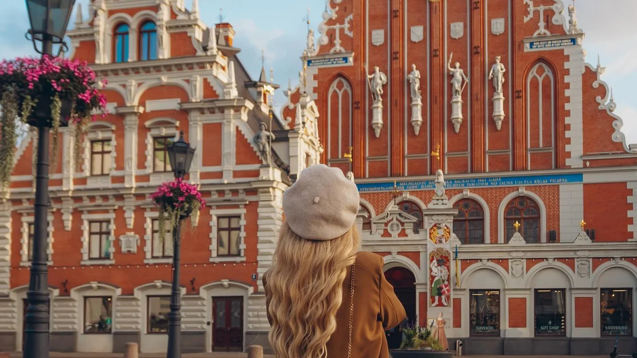 A woman with long blonde hair stands in Riga’s historic Old Town, gazing at the ornate red-and-white House of the Black Heads, a masterpiece of Gothic architecture, under a soft golden sky.