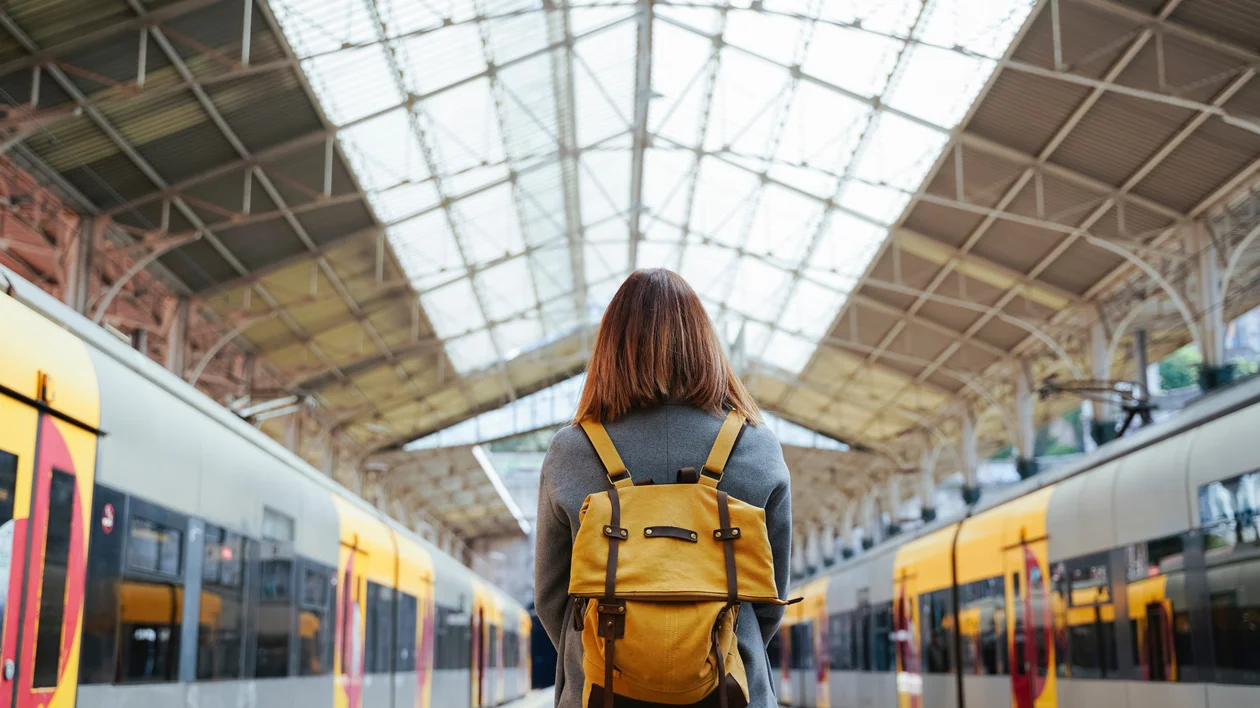A solo traveler with a mustard yellow backpack stands on a train platform beneath a grand, glass-roofed station, facing two sleek yellow and gray night trains ready for departure.