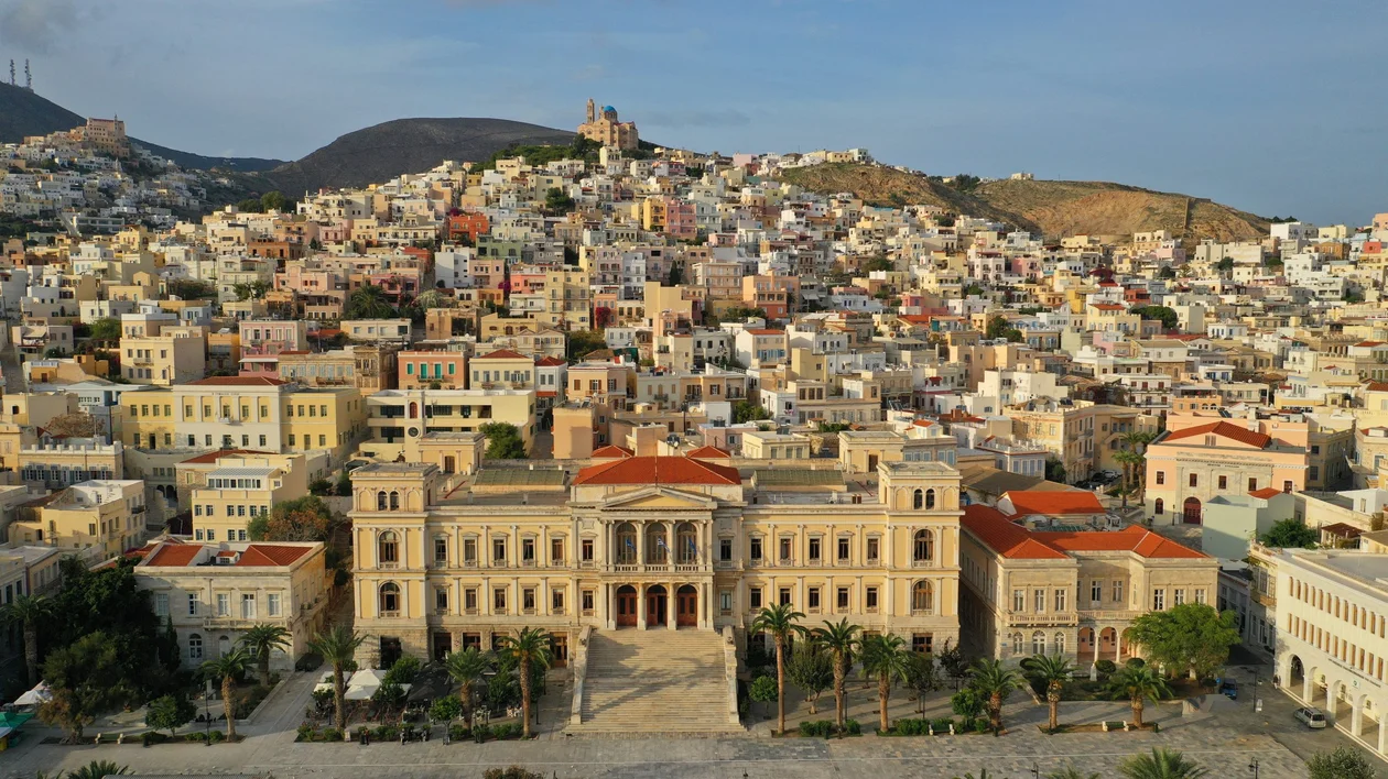 A panoramic view of Syros town with colorful houses cascading down the hillside, centered around a grand neoclassical building.