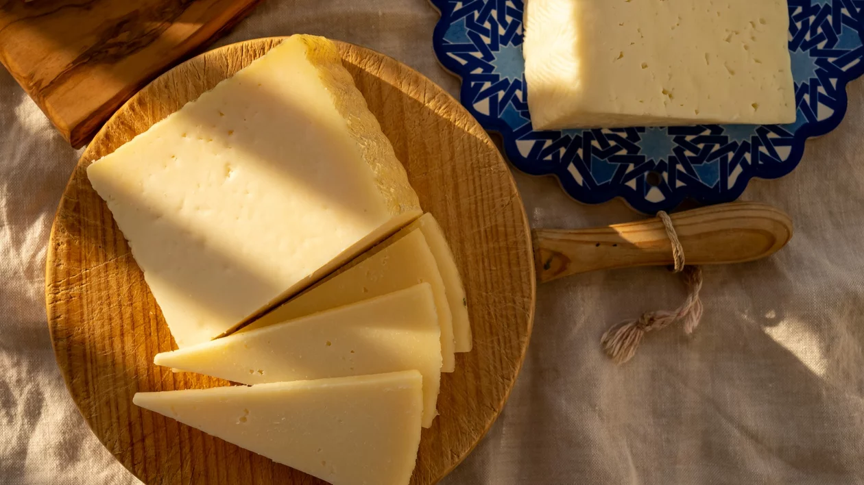 A cheesemaker holding a fresh wheel of Manchego cheese, surrounded by rustic cheesemaking tools in La Mancha, Spain.