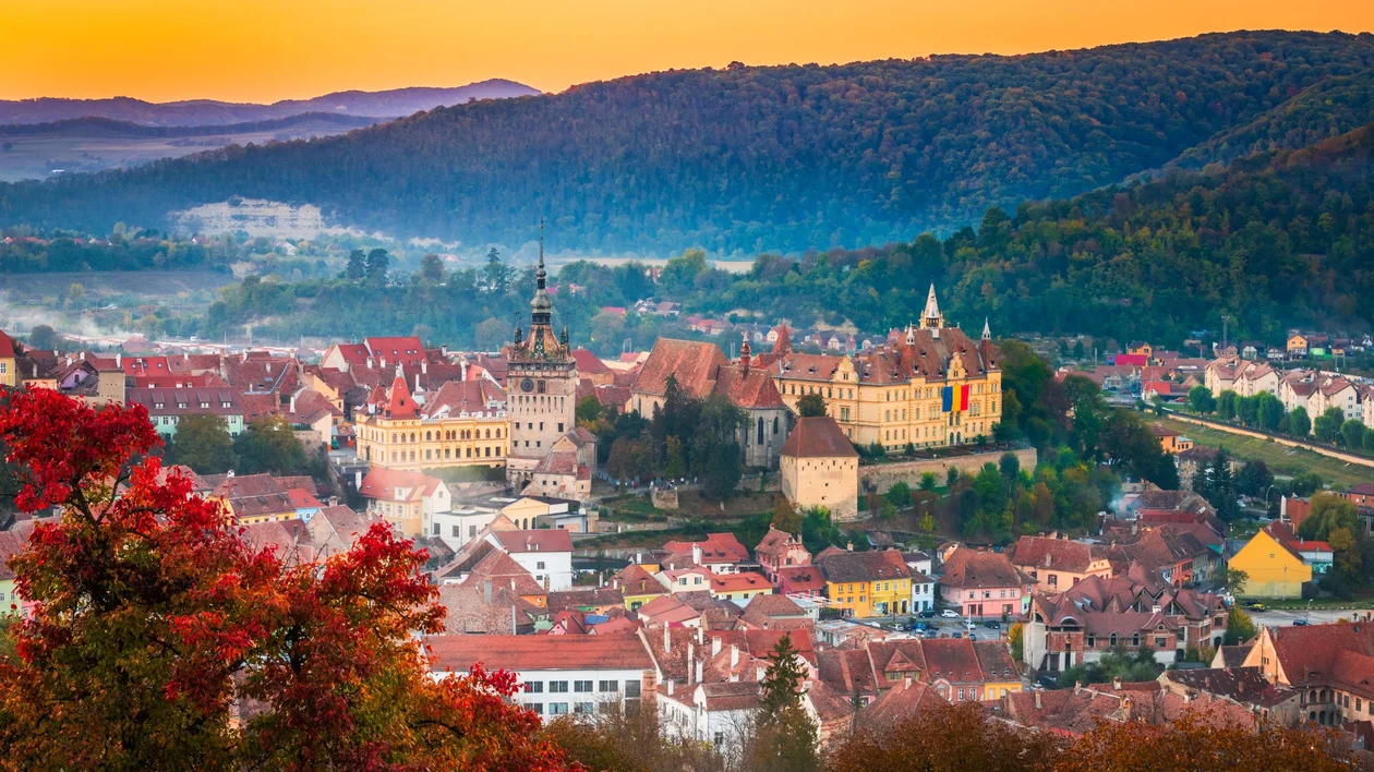 The medieval town of Sighisoara in autumn, with vibrant foliage framing historic buildings and a glowing sunset sky.