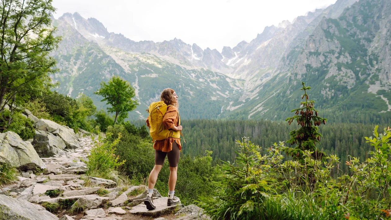A hiker walks along a scenic mountain trail in the Tatra Mountains, where the path itself forms the border between Poland and Slovakia. Snow-capped peaks, rugged slopes, and a deep blue alpine lake stretch into the distance under a bright, clear sky.