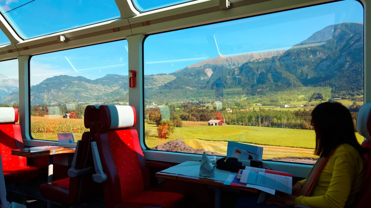 A woman relaxes in a panoramic train carriage, gazing out at sun-drenched alpine fields and mountains through large windows, as sunlight pours into the cozy red-seated cabin.