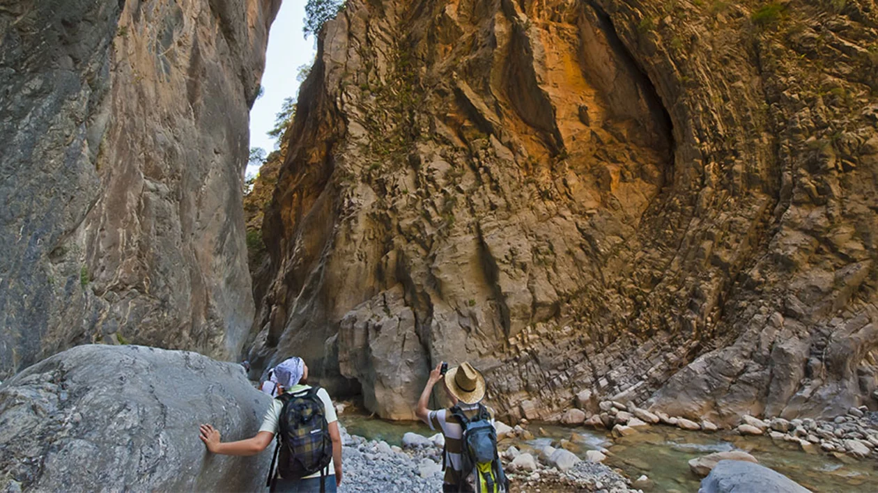 Two hikers explore a narrow canyon, capturing the towering rock walls and tranquil river below amidst lush greenery.