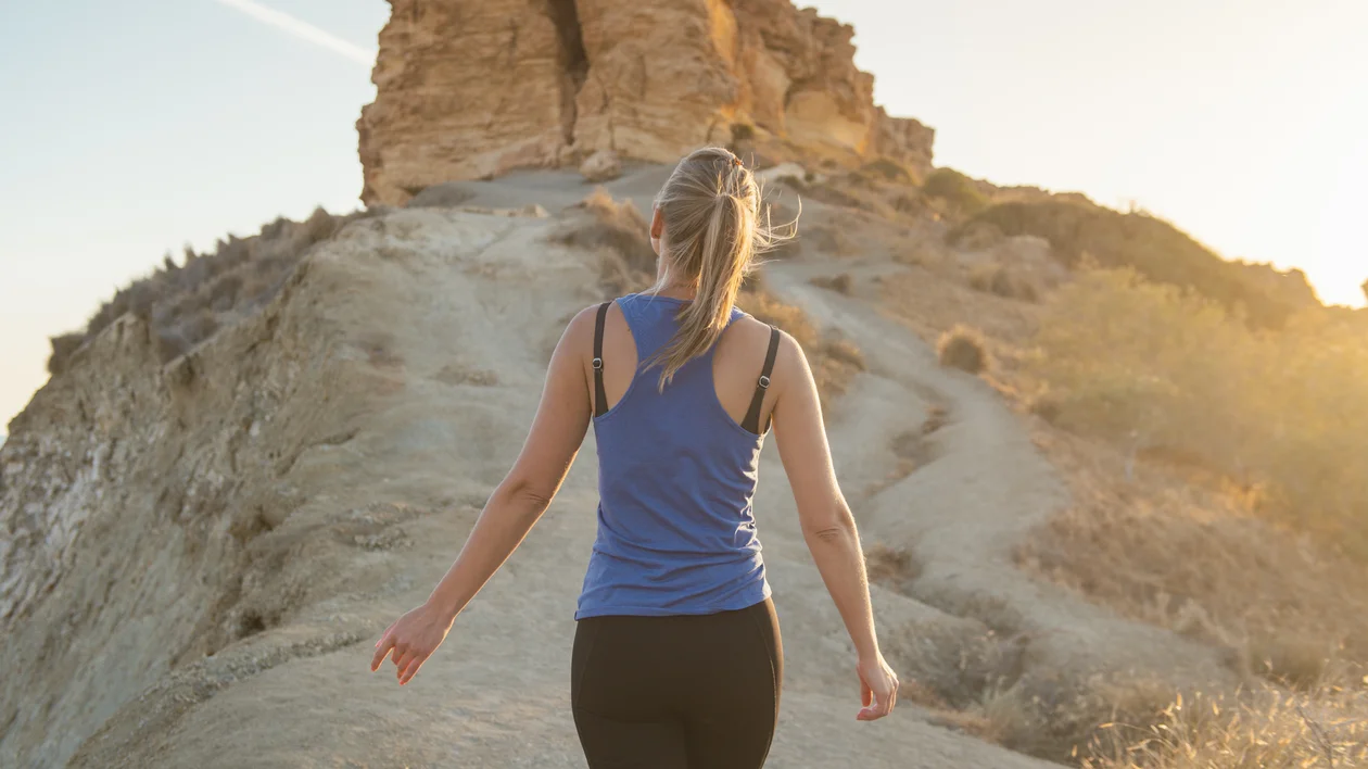 A woman walks along a narrow dirt trail through arid terrain, heading toward rocky outcrops under soft evening light.