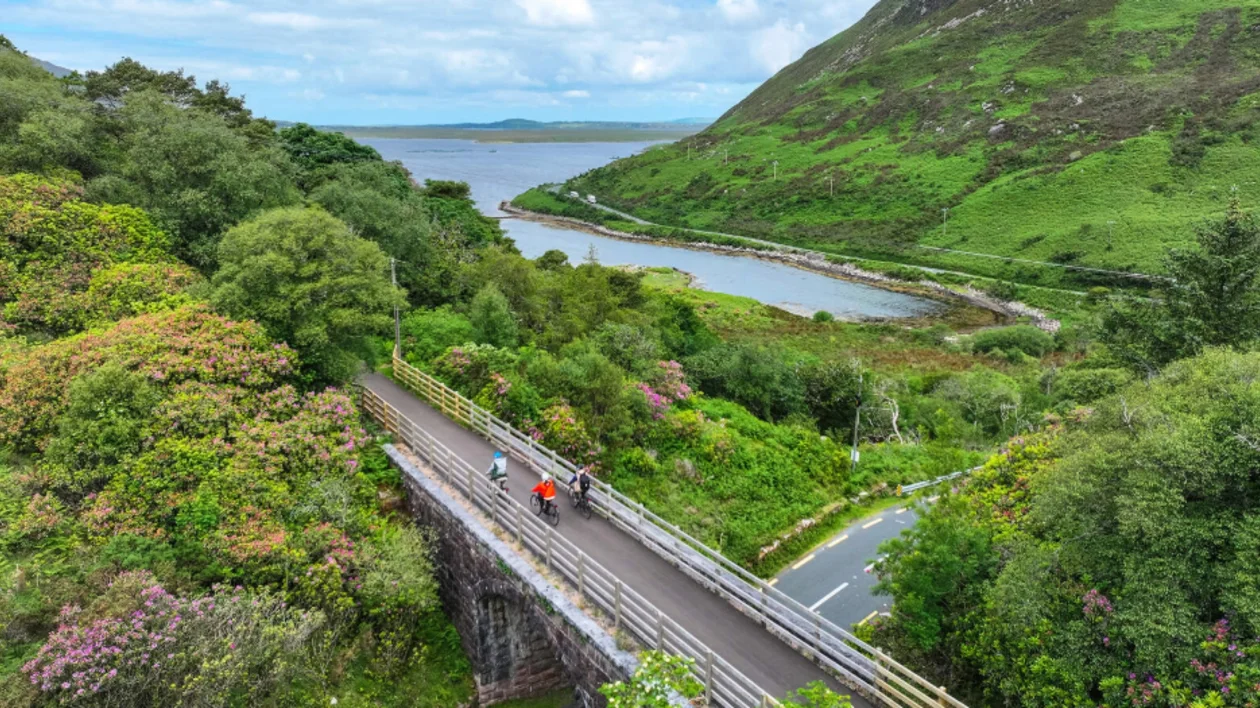 Two cyclists ride on a winding road beside vibrant greenery and a serene river, with a hilly landscape under a cloudy sky.
