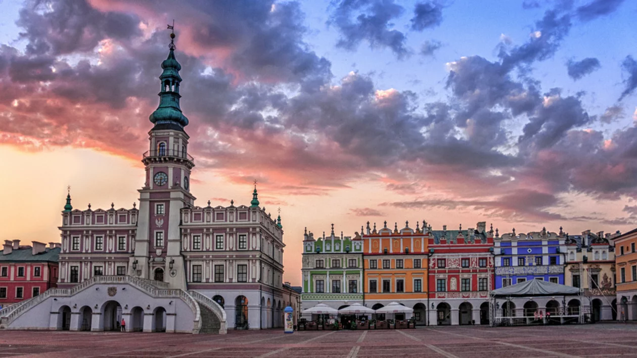 Twilight view of Zamość Market Square, featuring colorful historic buildings and a prominent clock tower amidst a dramatic sky.