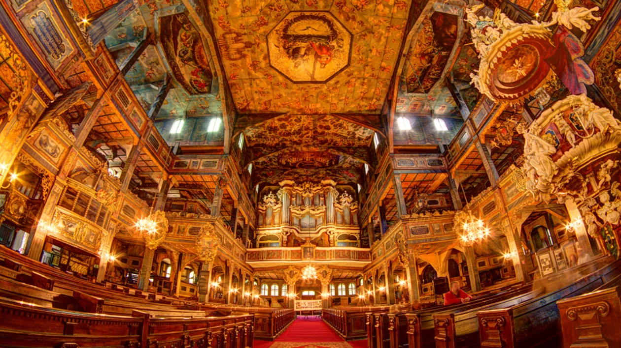 Interior of an ornate church featuring intricate paintings, wooden pews, chandeliers, and a grand organ, bathed in warm light.