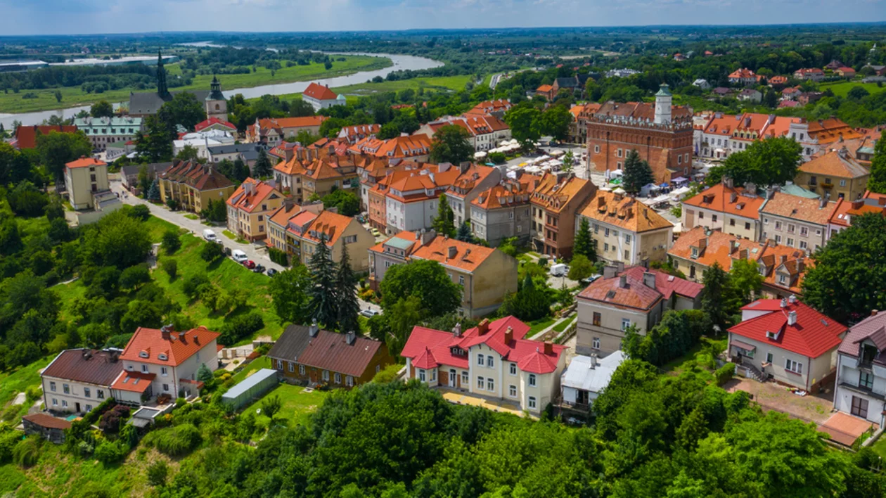 Aerial view of a picturesque town with red-roofed houses, green trees, and a river in the background under a bright blue sky.