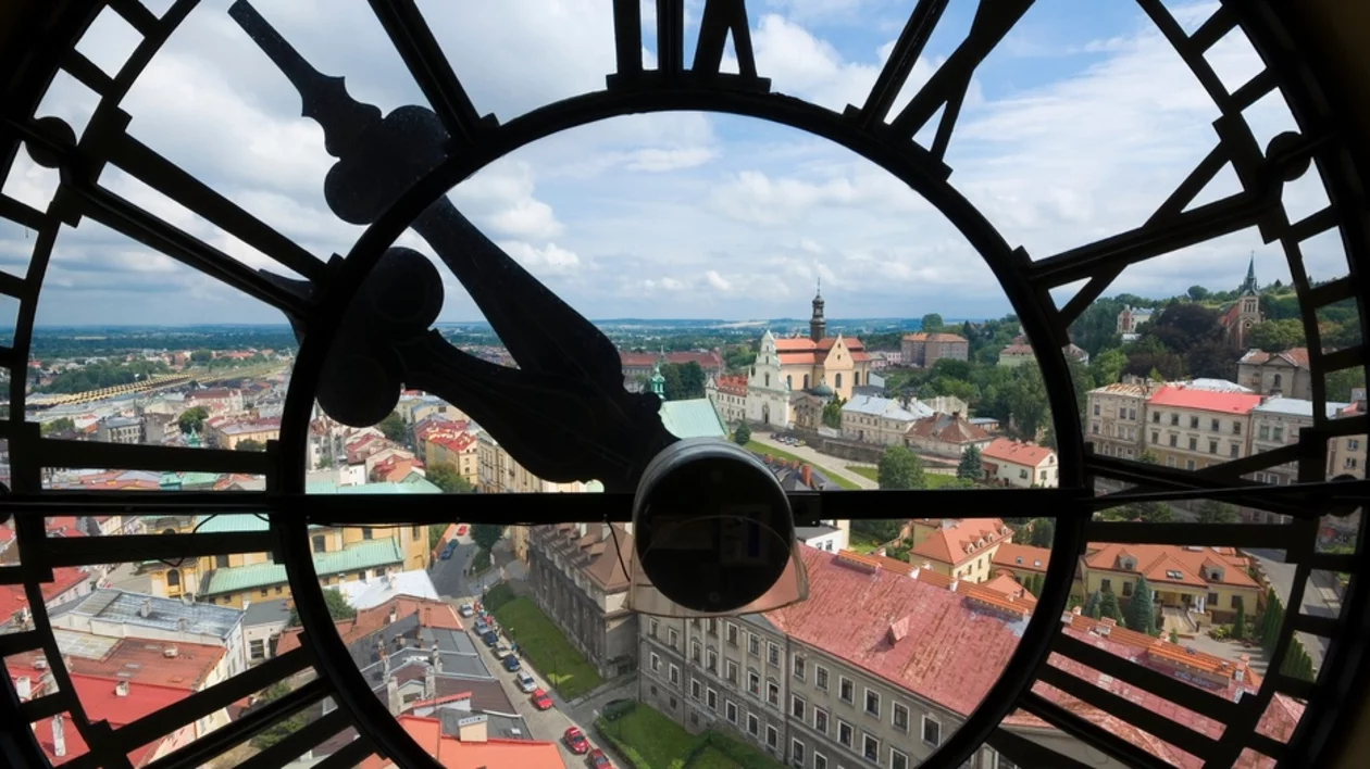 View through a large clock face, revealing a picturesque town with red-roofed buildings and lush green hills under a cloudy sky.