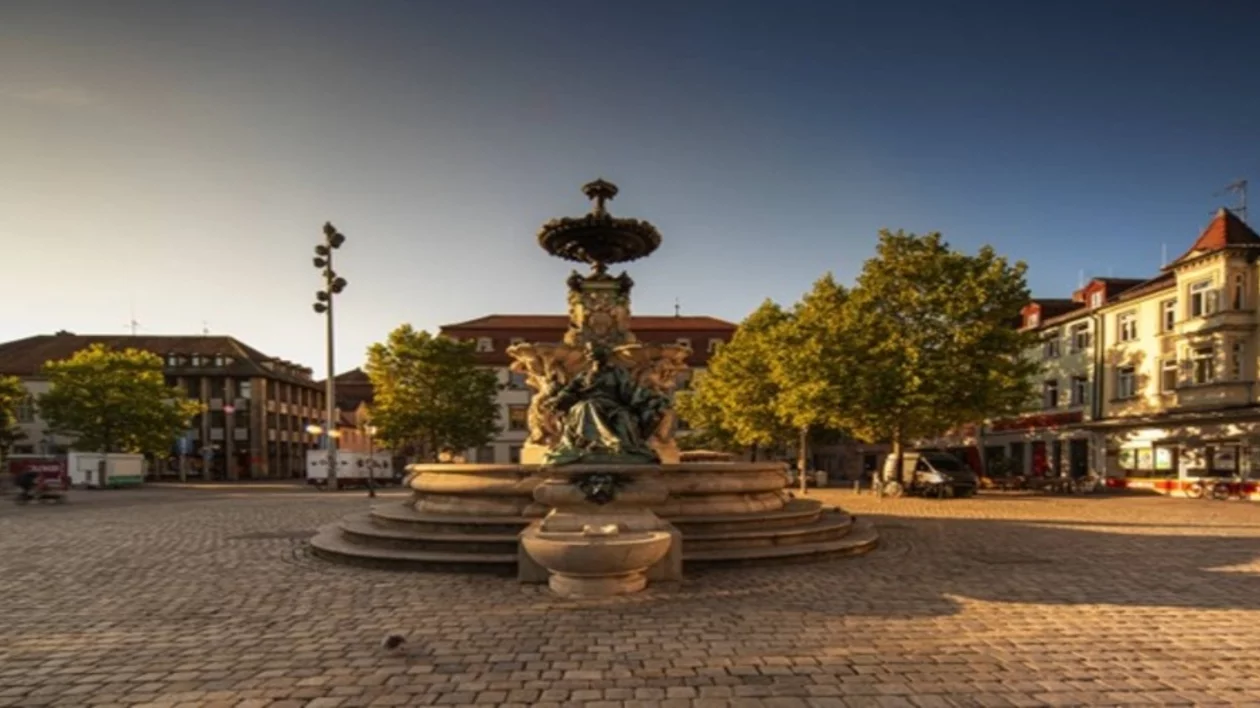 A historic fountain surrounded by trees and buildings, set in a sunny, open square with cobblestone pavement.