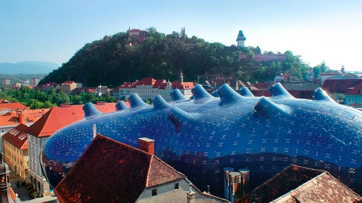 A futuristic blue building, the Kunsthaus Graz, contrasts with traditional rooftops and a green hillside in Graz, Austria.