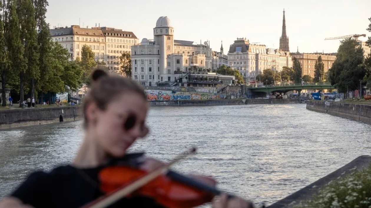 A riverside scene featuring a person playing the violin, with buildings, trees, and a bridge in the background during sunset.