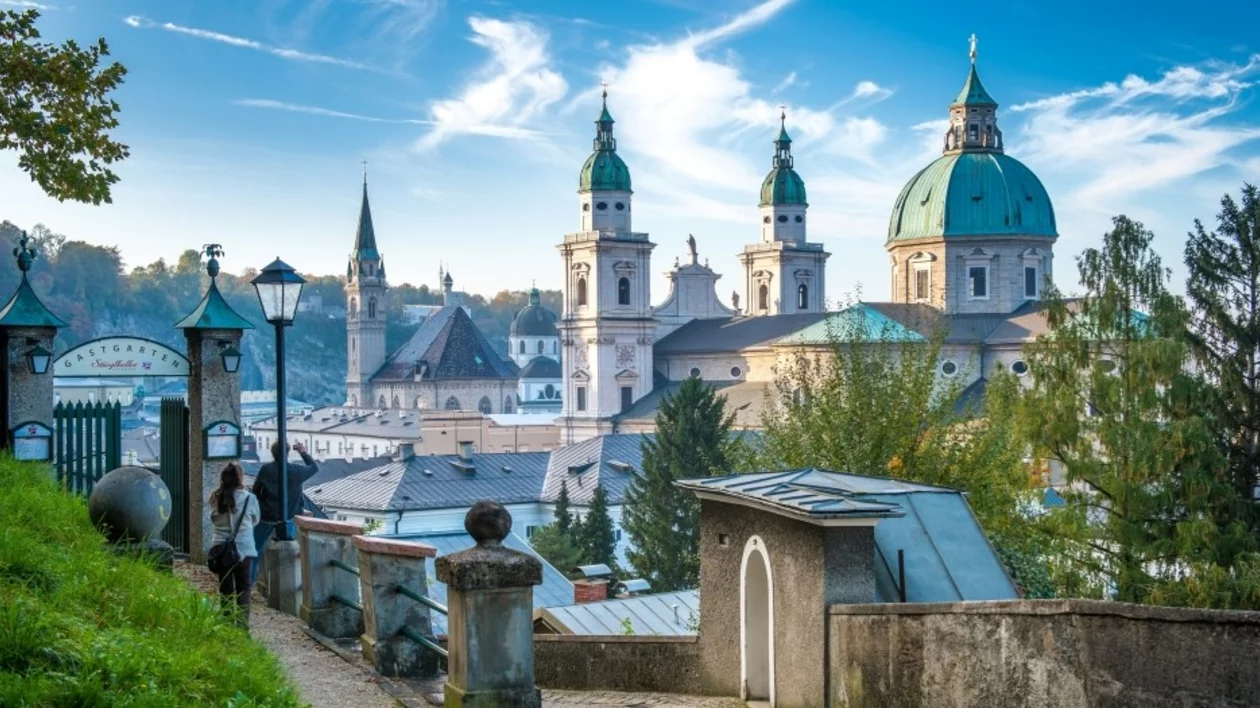 A scenic view of a historic city with green-domed churches and a clear blue sky, framed by trees and street lamps.