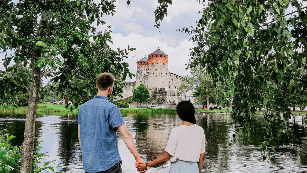 Couple holding hands by the lake, looking at the medieval Olavinlinna Castle in Savonlinna, Finland.